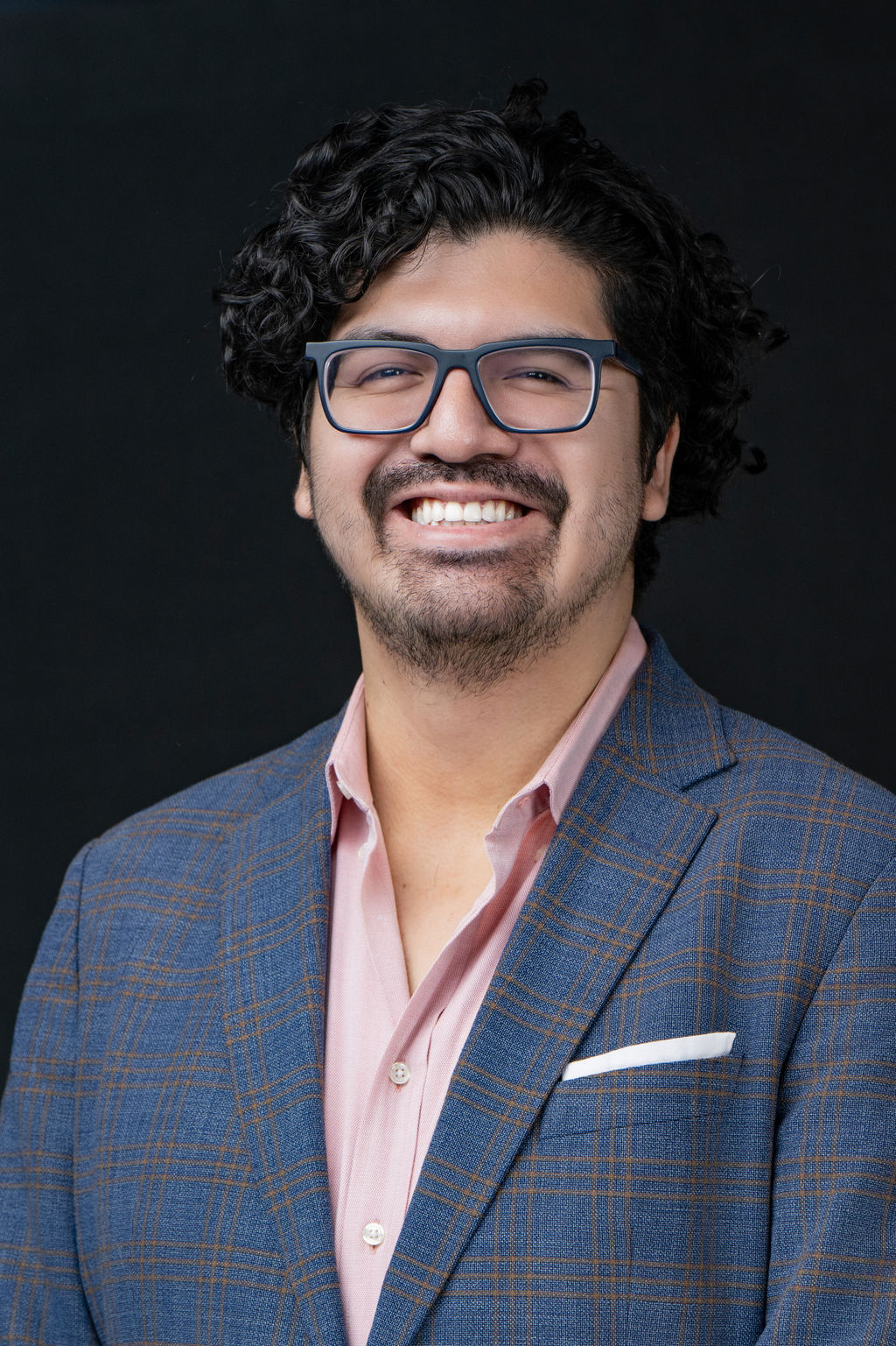 Studio headshot of a smiling man in a blue plaid blazer and pink shirt