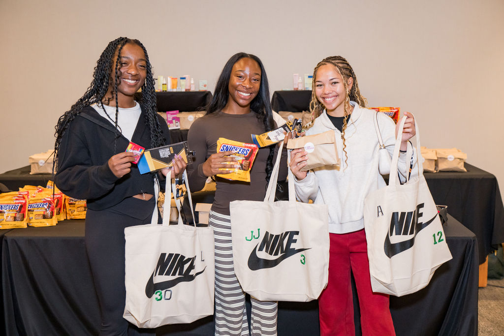 Three student athletes smiling with Nike tote bags at the BSA Summit sponsor table