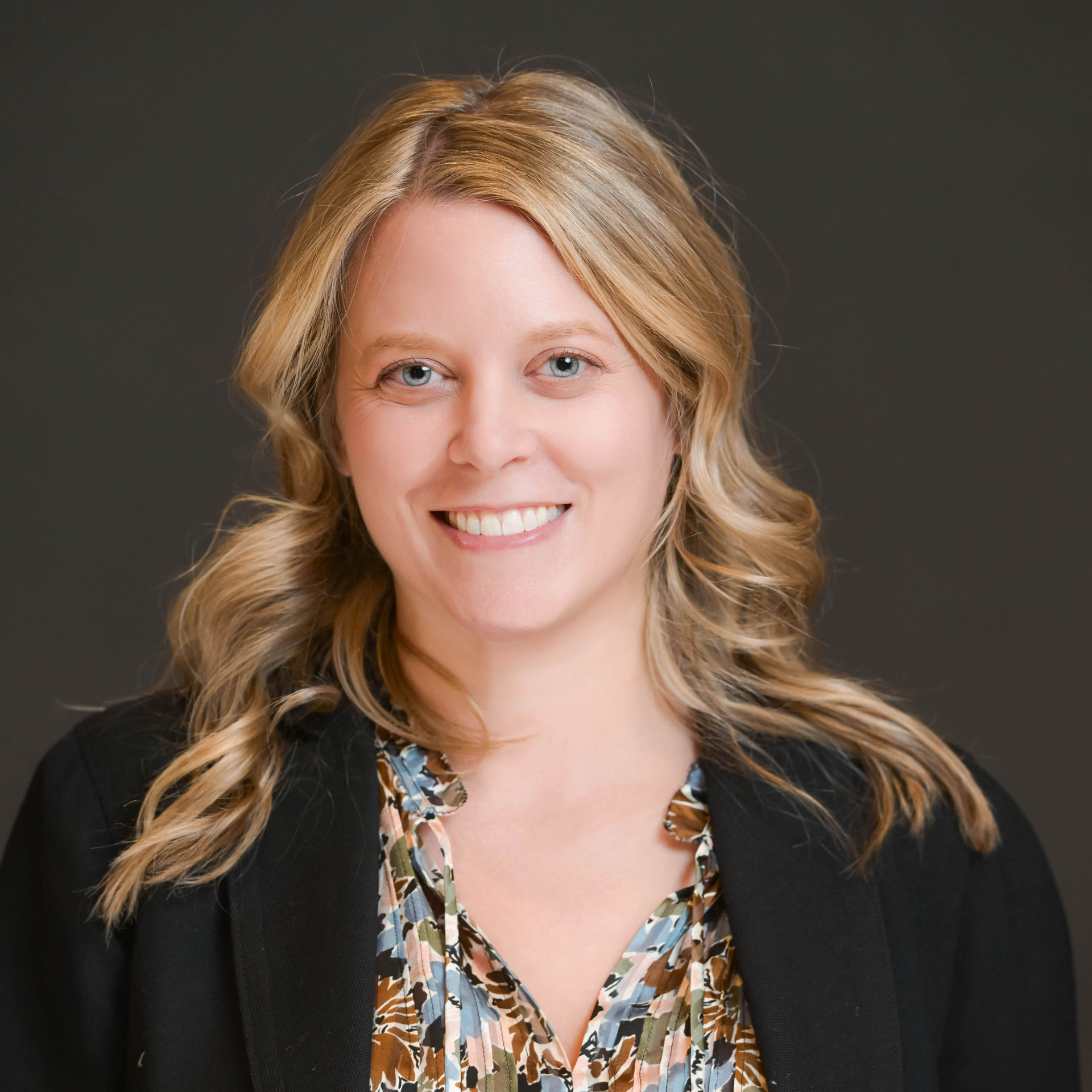 Studio headshot of a professional woman in a black blazer on a grey backdrop