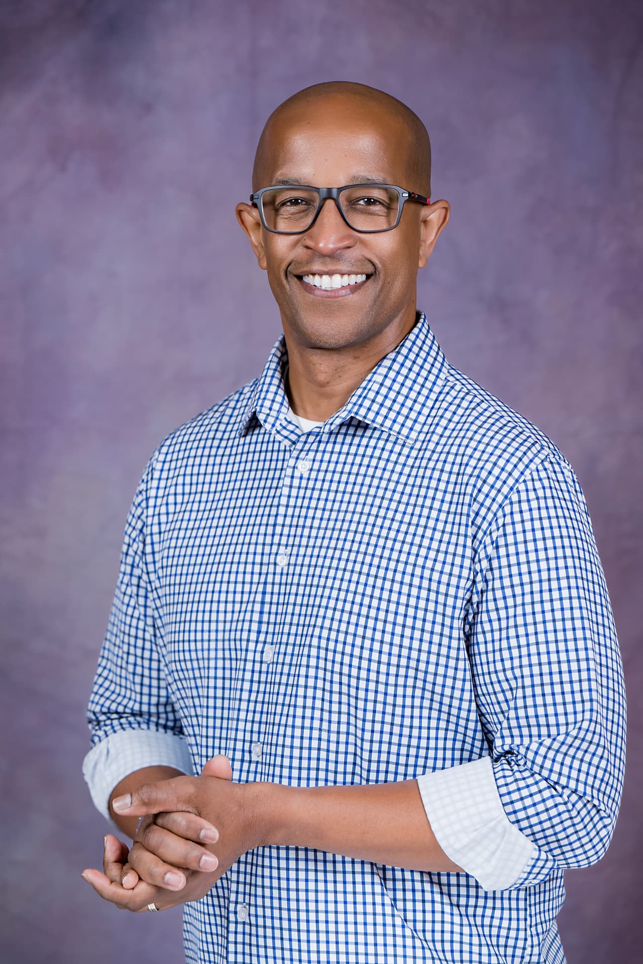 Studio portrait of a smiling man in a blue checked shirt and glasses