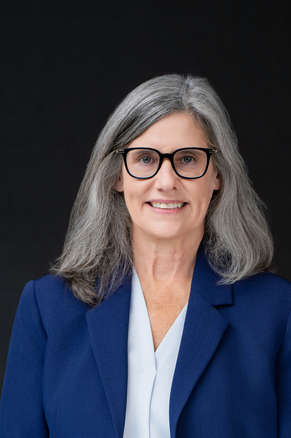 Studio headshot of a woman with silver hair and glasses in a navy blazer