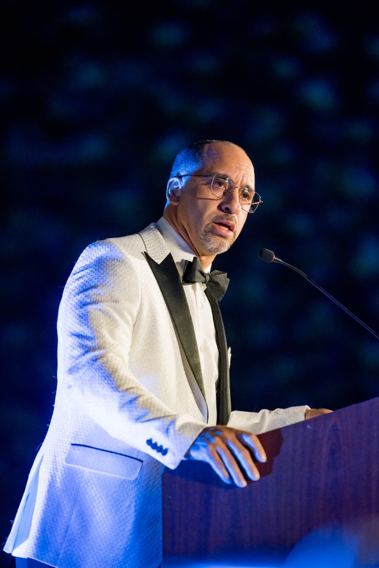 Speaker in a white tuxedo addressing guests at a black-tie gala