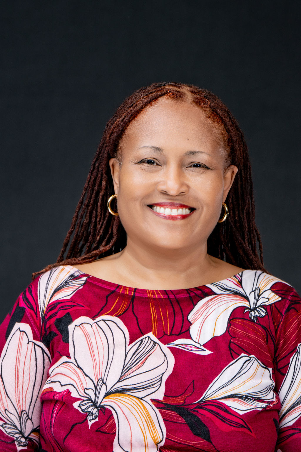 Studio headshot of a smiling woman with locs in a floral burgundy top