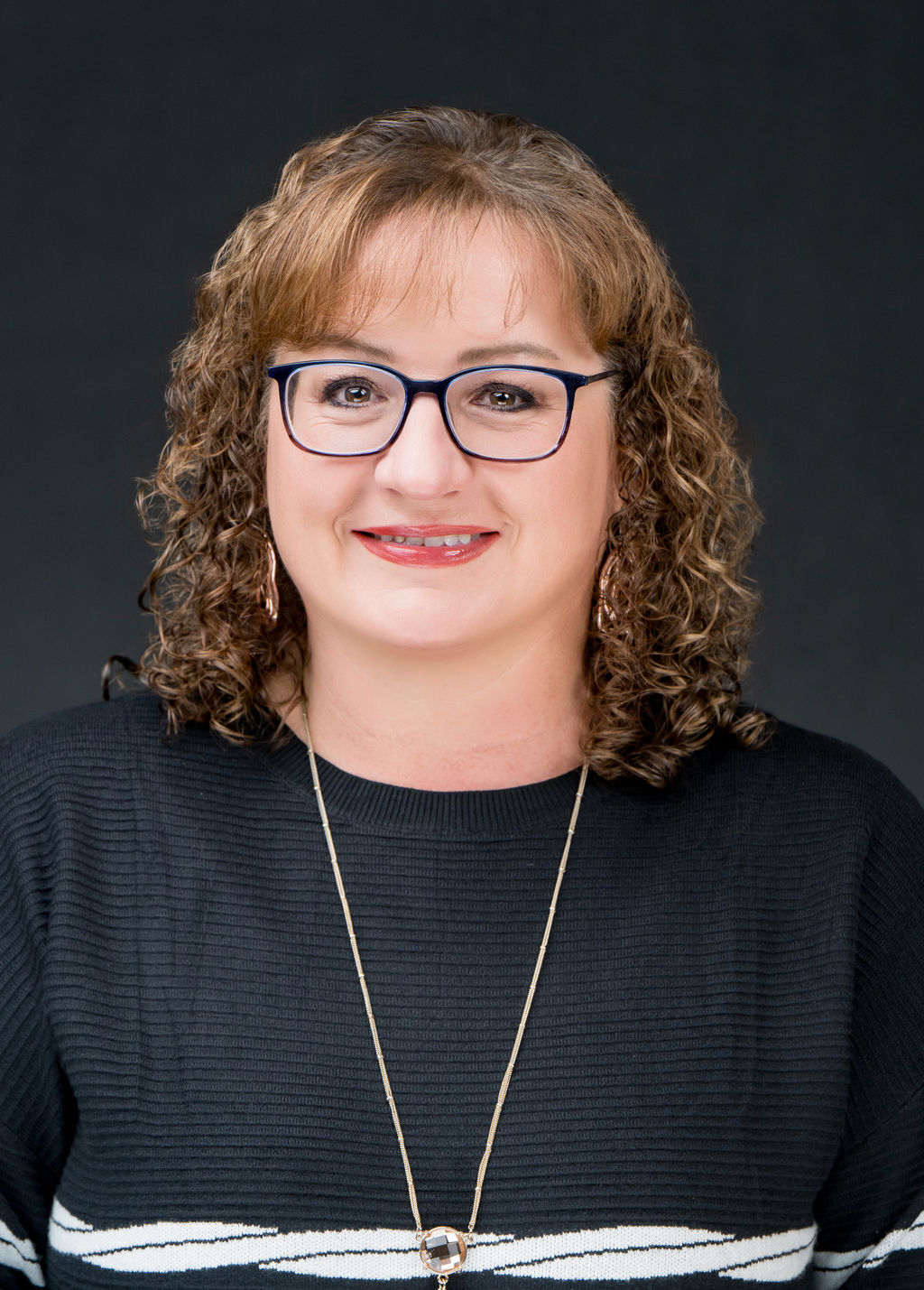 Studio headshot of a smiling woman with curly hair and navy glasses in a black sweater
