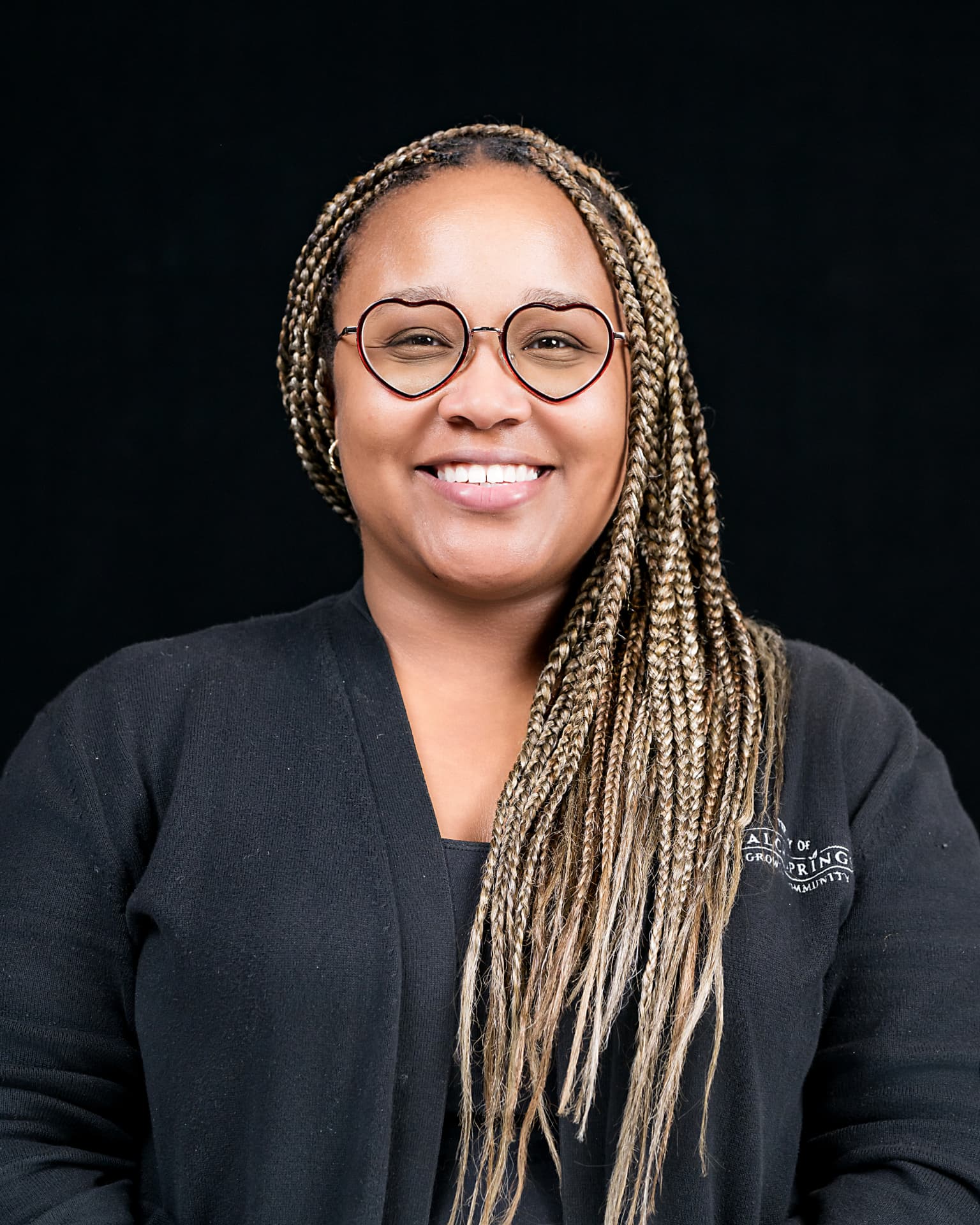 Studio headshot of a smiling professional with braided hair and round glasses
