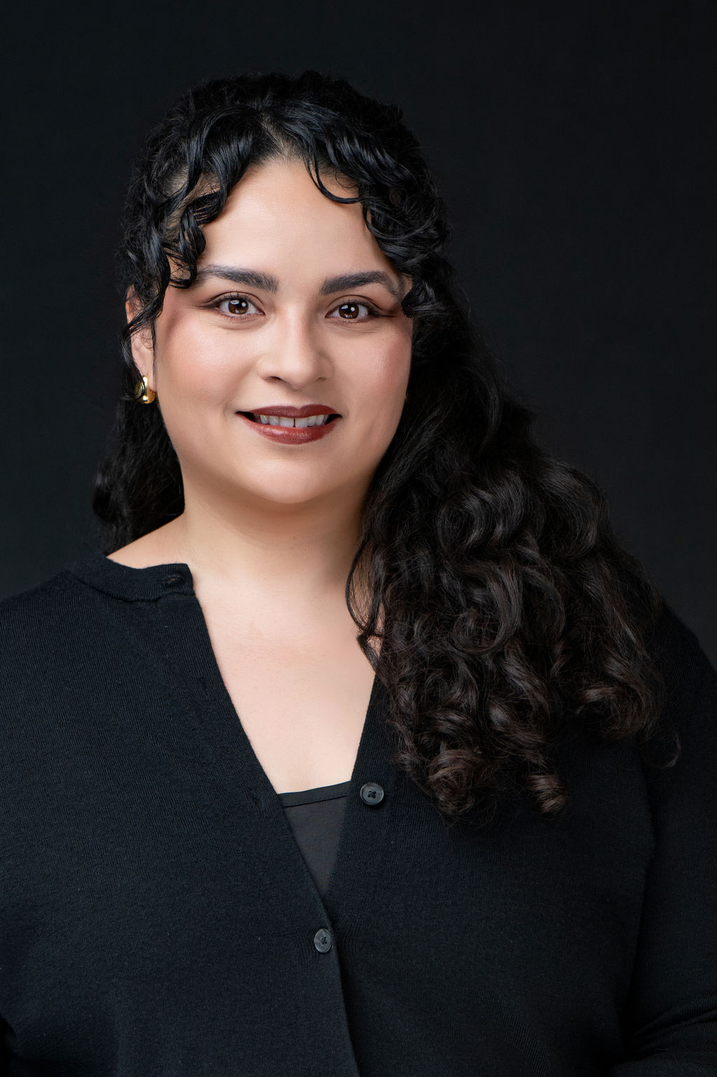 Studio headshot of a smiling woman with curly dark hair in a black cardigan