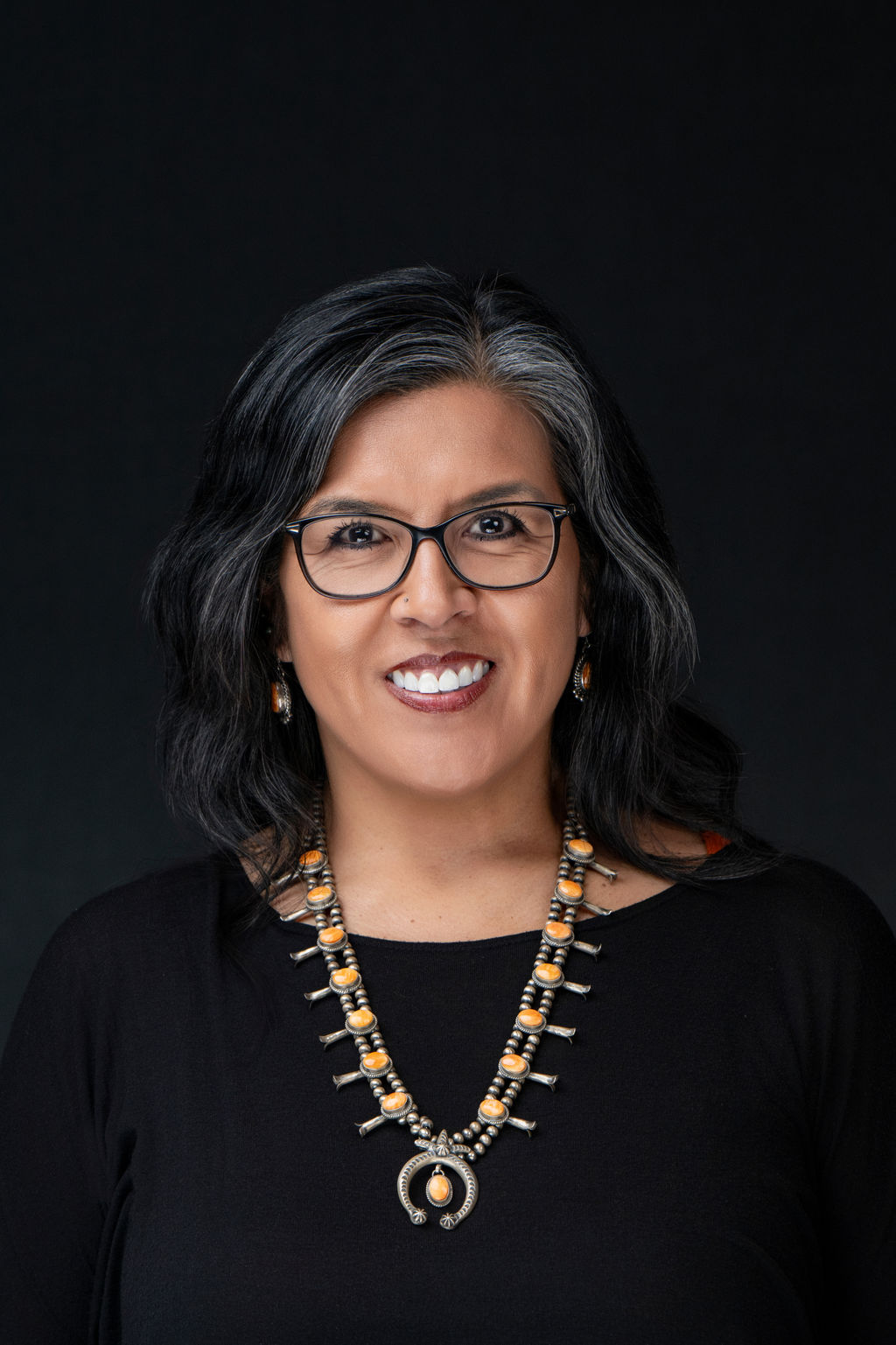 Studio headshot of a smiling woman with glasses and a turquoise statement necklace