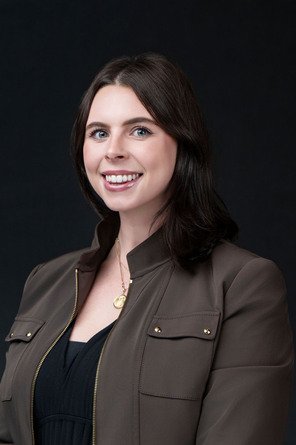 Studio headshot of a smiling woman with dark hair in an olive utility jacket