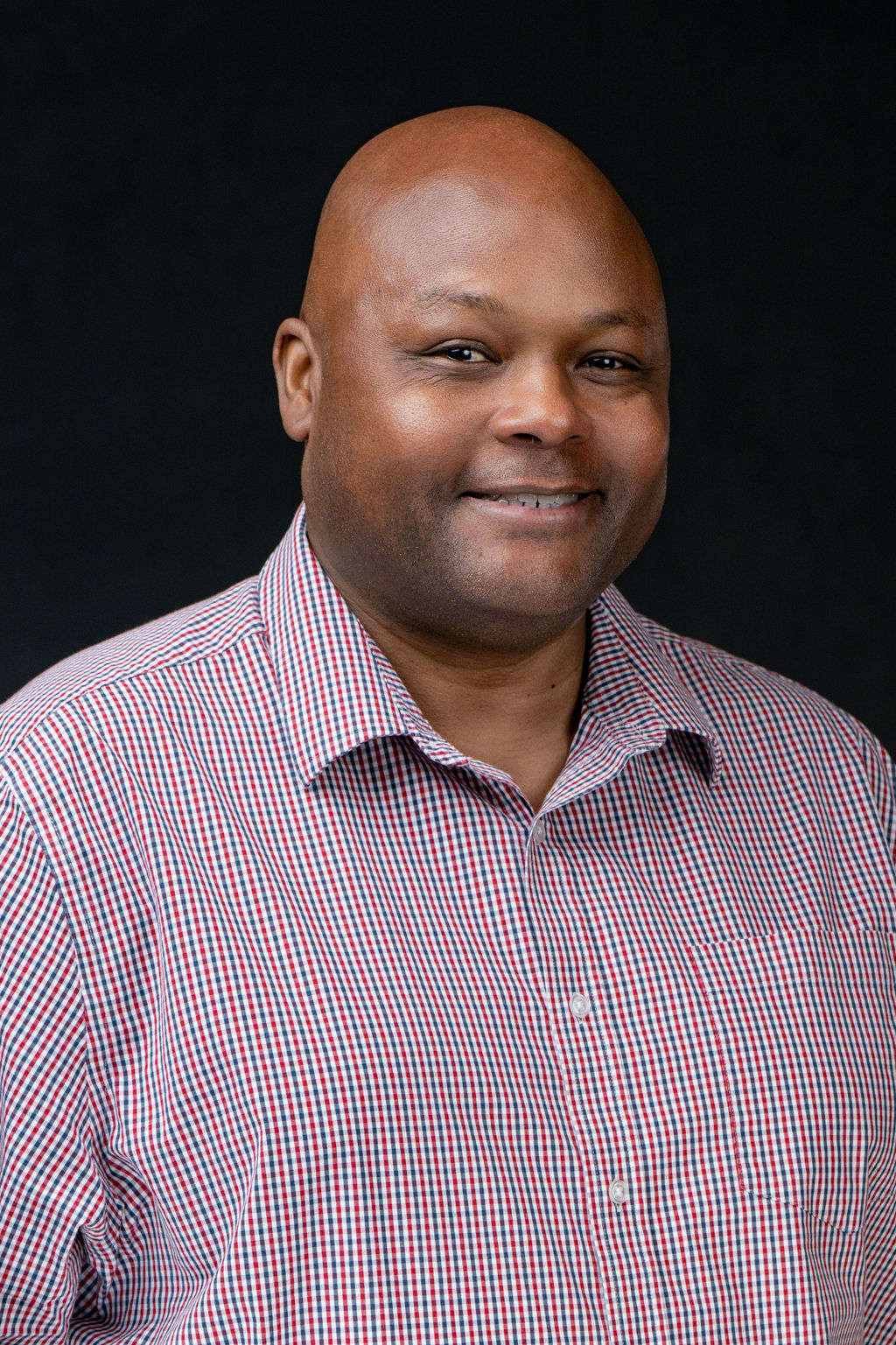 Studio headshot of a smiling man in a red and navy checked shirt