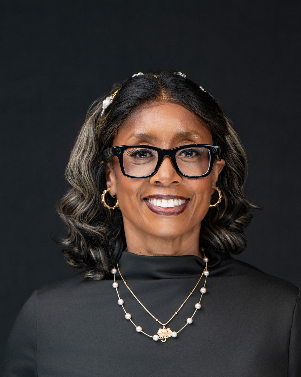 Studio headshot of a smiling woman with glasses and pearl necklace in black