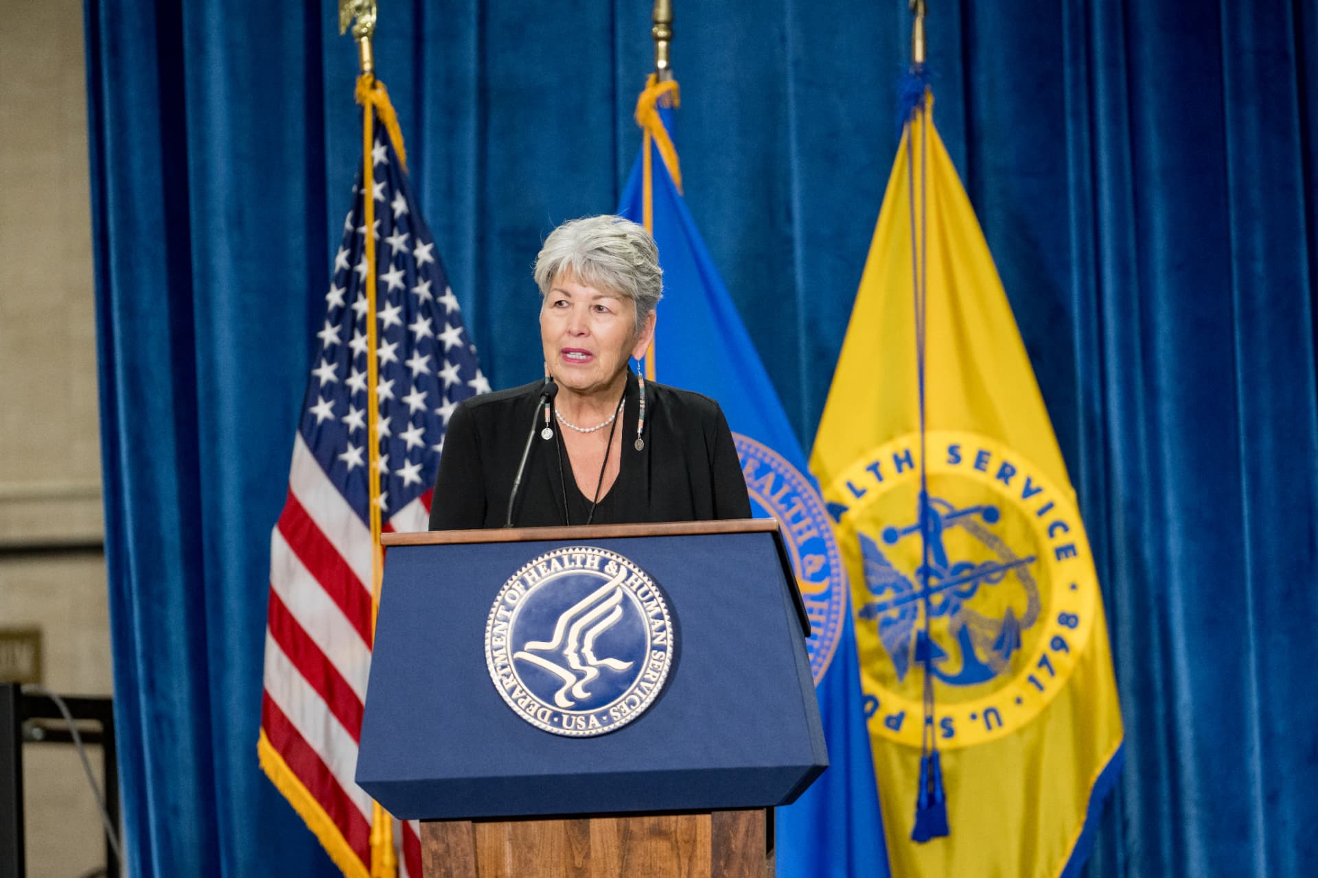 Speaker at a federal health agency podium flanked by the American flag and service flags