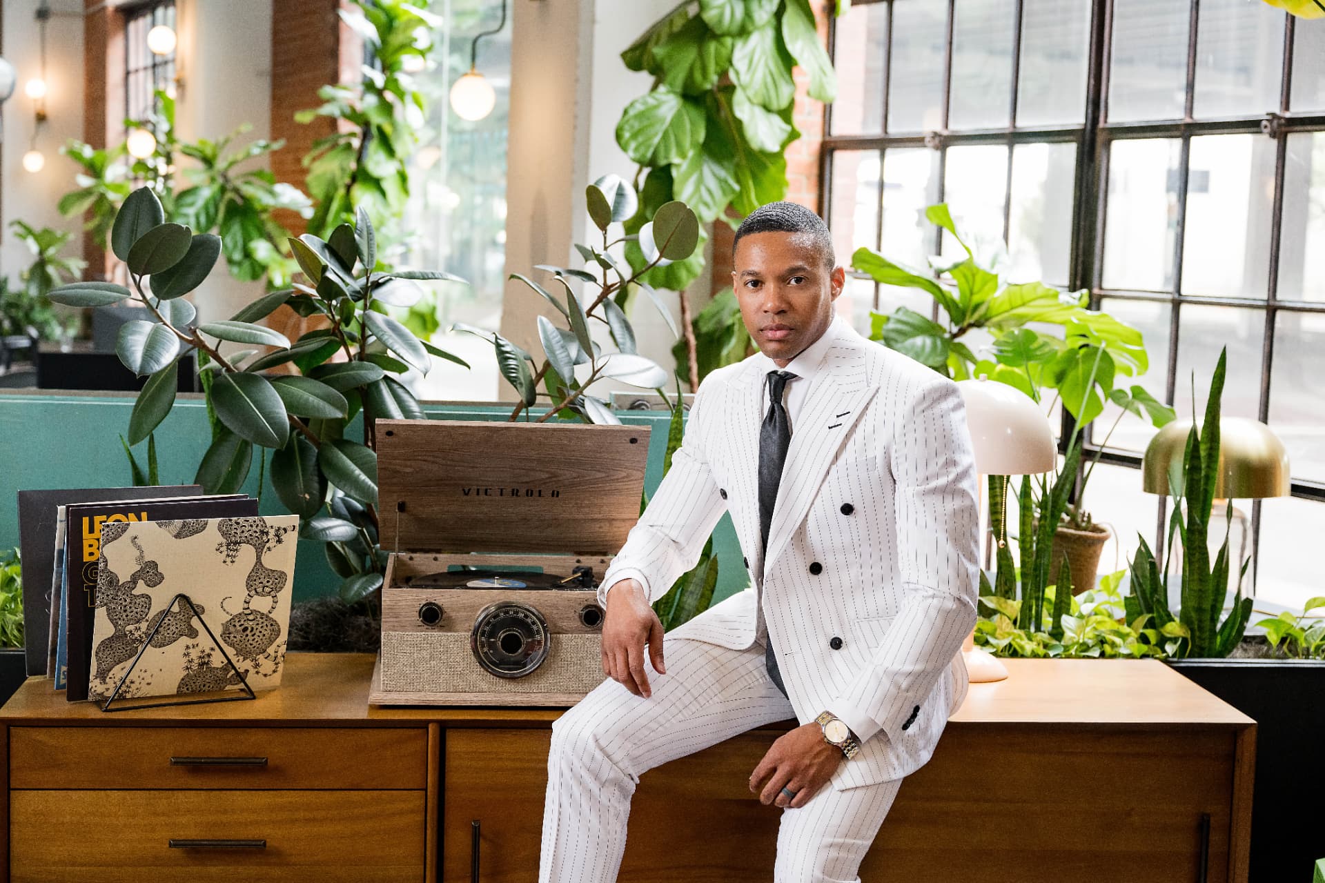 Man in a white pinstripe suit seated beside a record player in a plant-filled loft