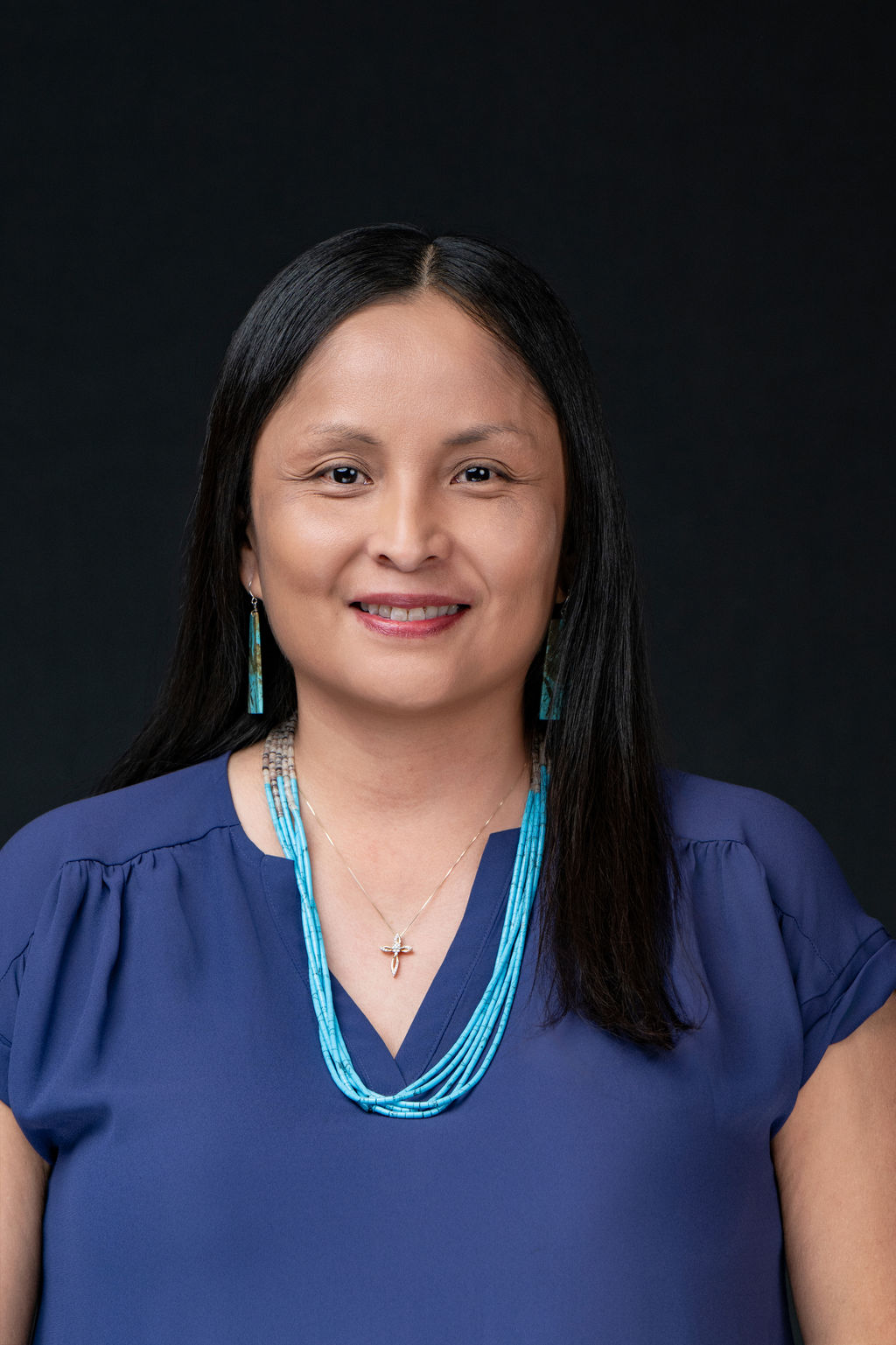 Studio headshot of a smiling woman in a navy blouse with turquoise jewelry