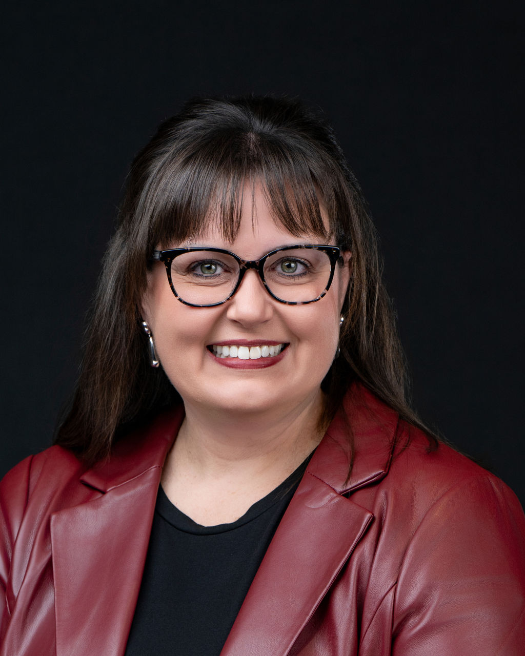 Studio headshot of a smiling woman with glasses in a burgundy leather blazer