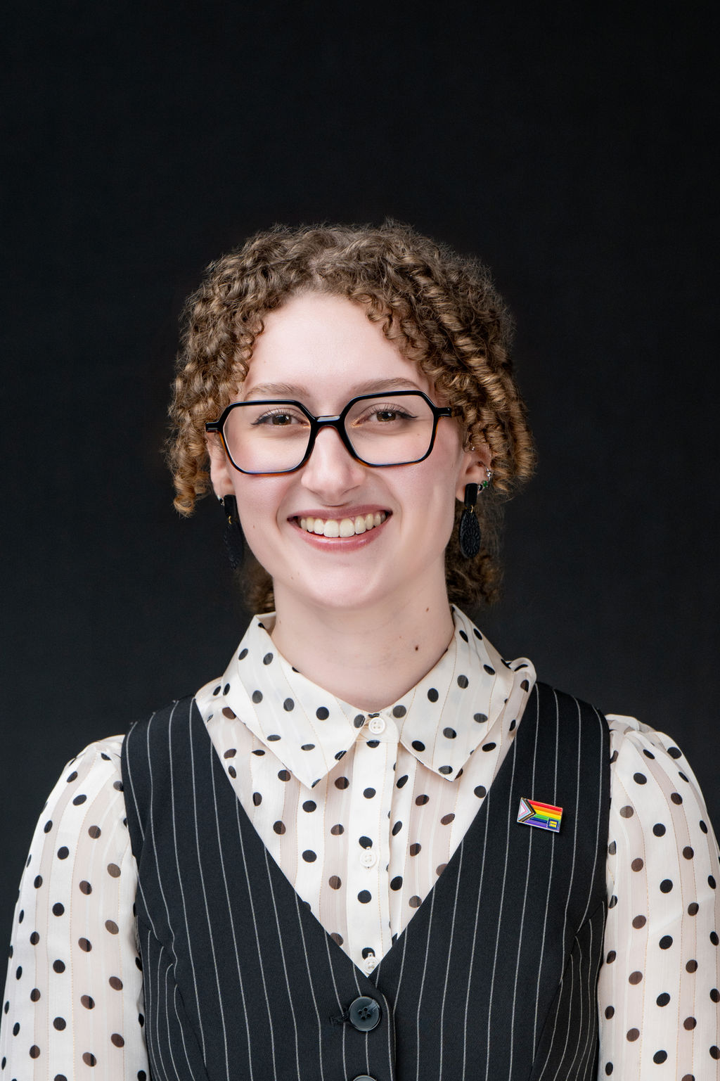 Studio headshot of a smiling person with curly hair, glasses and a pinstripe vest