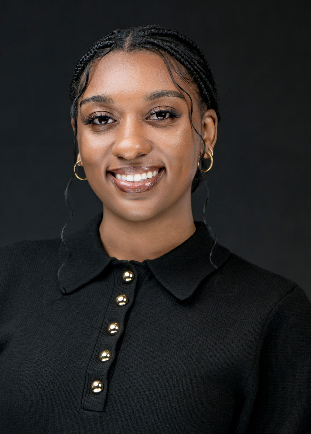 Studio headshot of a smiling woman with braids in a black collared sweater