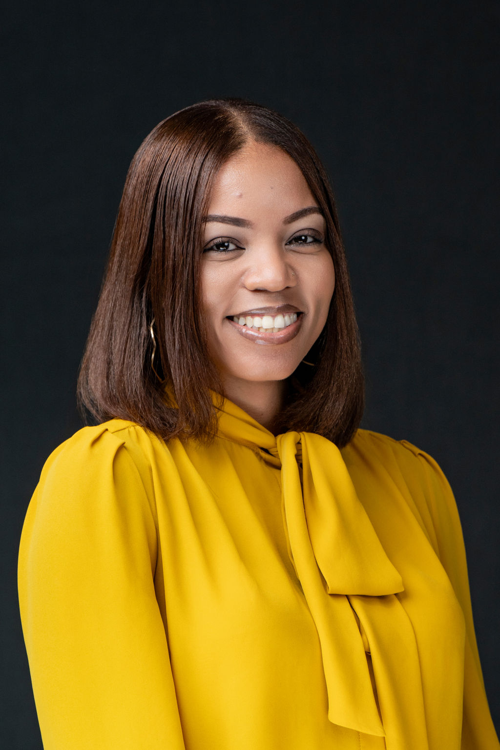Studio headshot of a smiling woman in a golden yellow tie-neck blouse