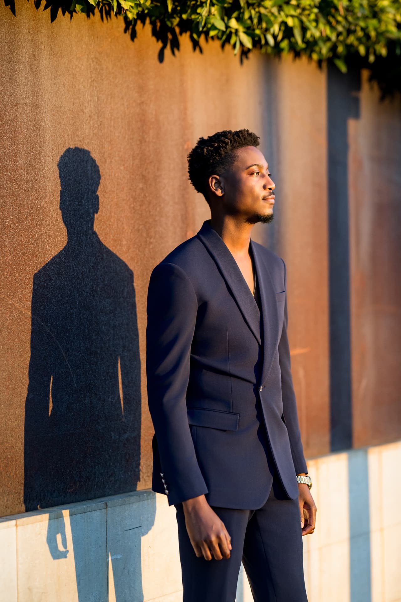 Model in a navy suit casting a shadow on a sunlit corten steel wall